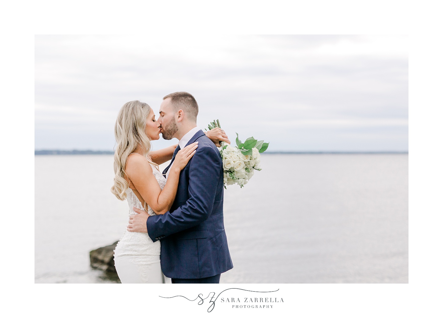 bride and groom kiss standing on rocks in front of Narragansett Bay