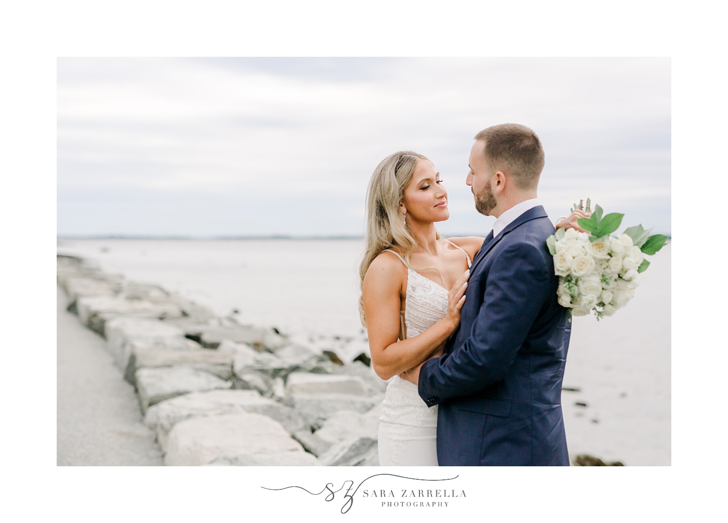 bride looks up at groom hugging on rock formation near Narragansett Bay