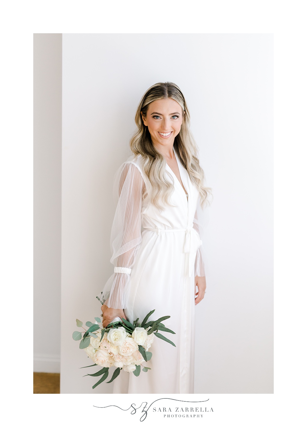 bride poses in suite at Belle Mer Island House in white lace robe holding bouquet 