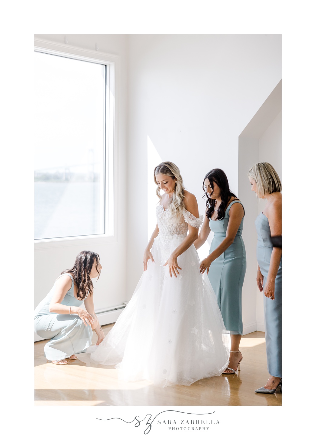 bridesmaids and mother help bride into wedding gown in front of window in bridal suite at Belle Mer Island House