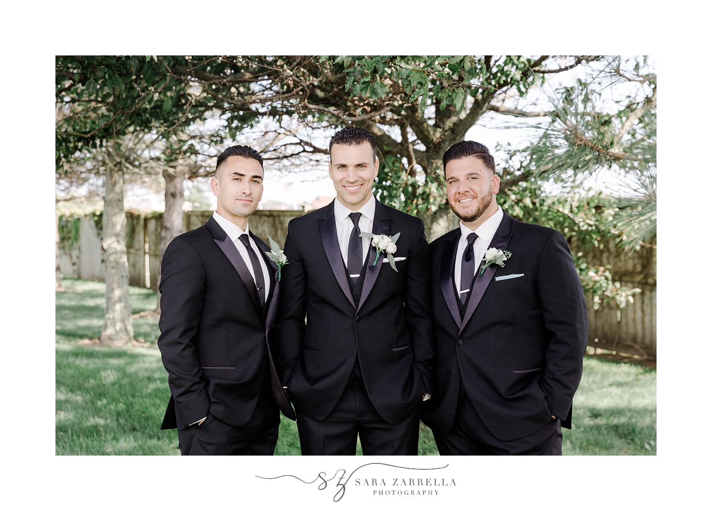 groom poses with two groomsmen in black suits on lawn at Belle Mer Island House