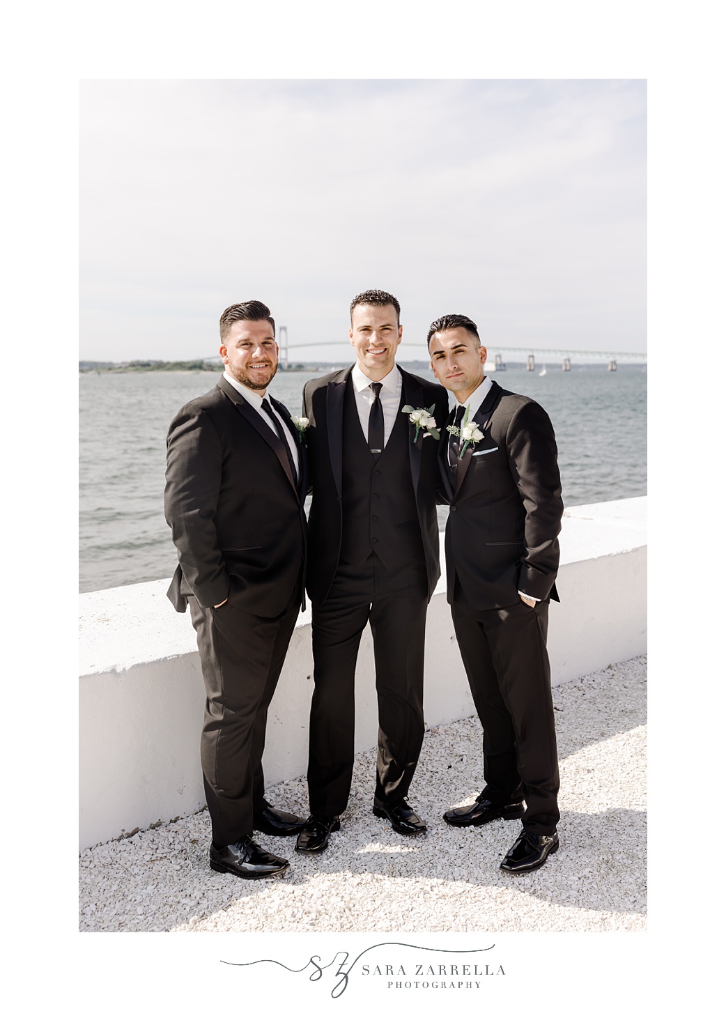 groom and two groomsmen in black suits pose alongside white wall