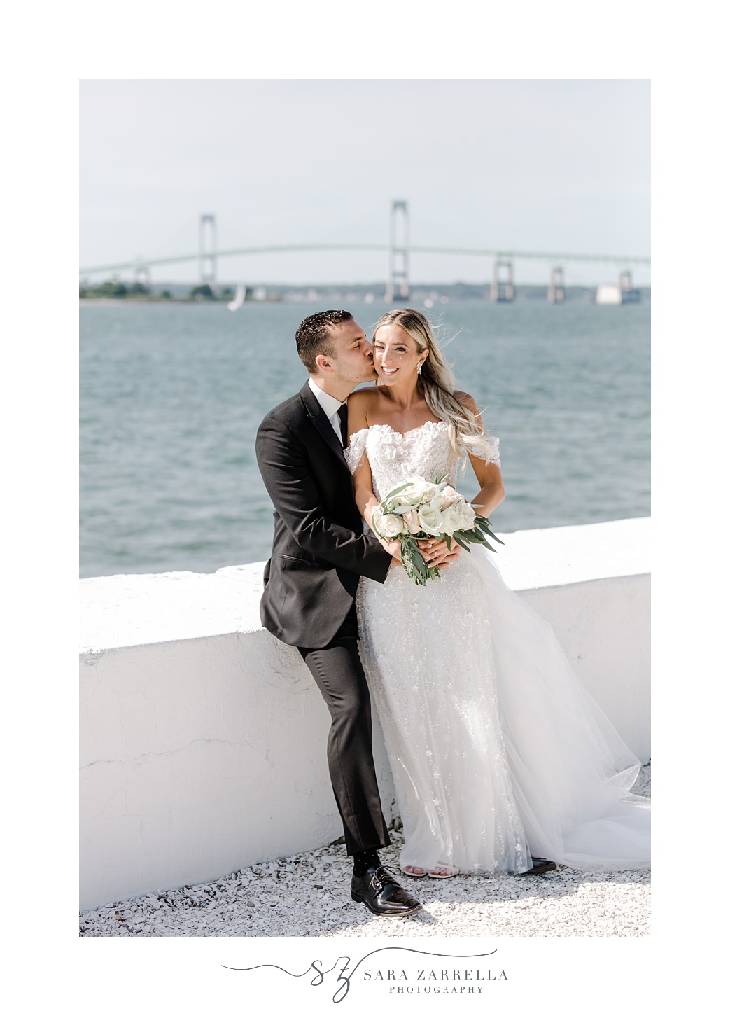 groom hugs bride to him as he leans on white wall of Belle Mer Island House