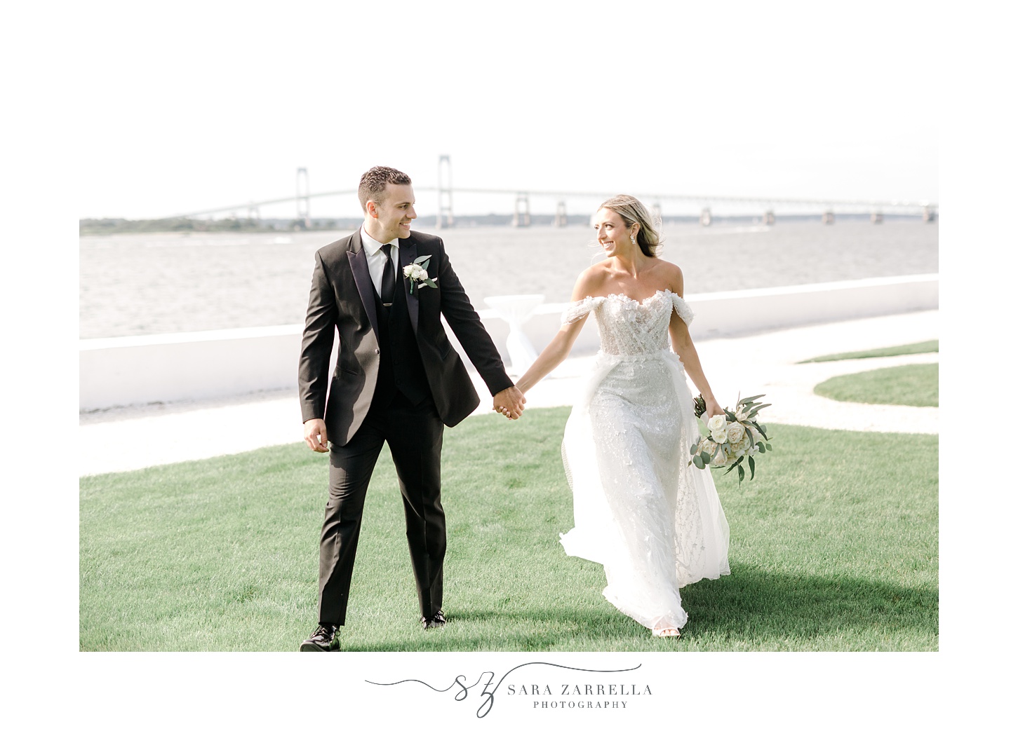 bride and groom hold hands walking away from Newport Bridge 