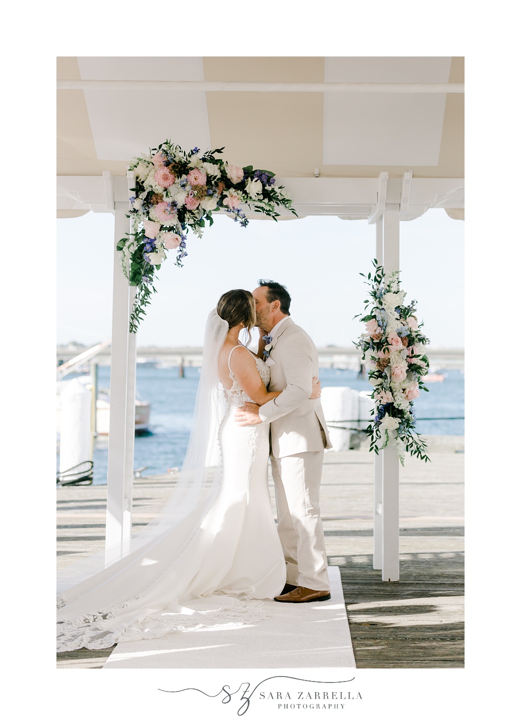 newlyweds kiss under white arbor with pink flowers during waterfront wedding ceremony at Regatta Place