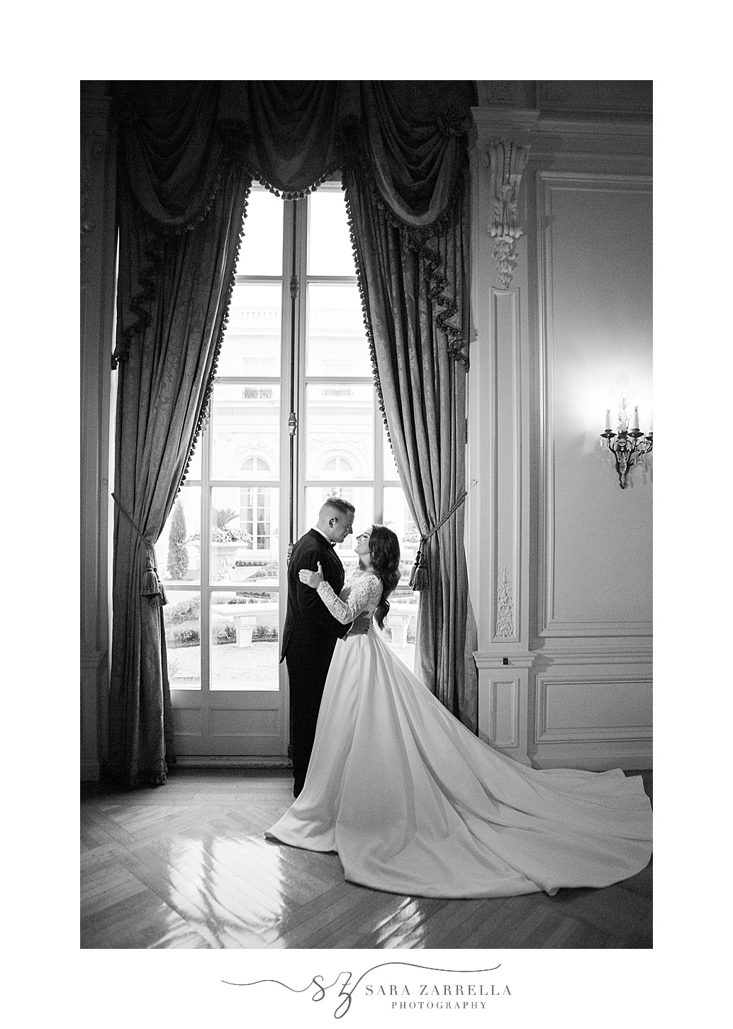 newlyweds hug in front of windows with bride's skirt draped behind her