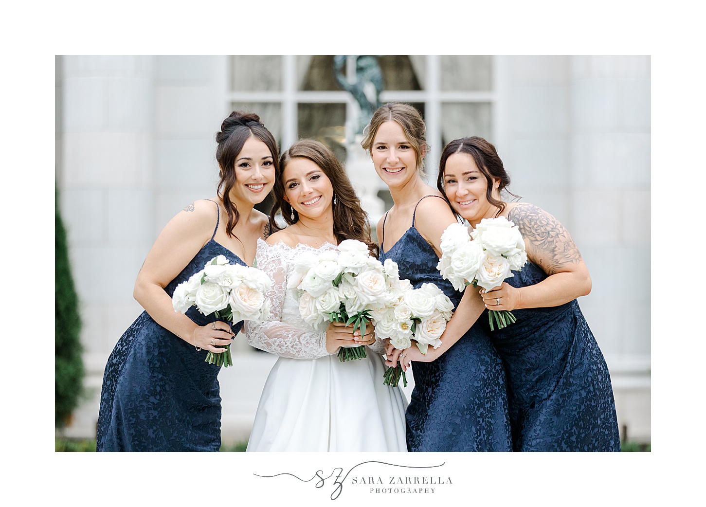 bride poses with bridesmaids in navy gowns with white flowers outside Rosecliff Mansion