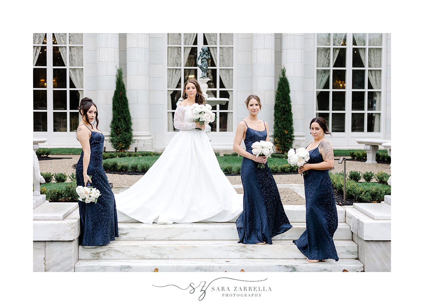 bride poses on steps outside Rosecliff Mansion with bridesmaids in navy gowns