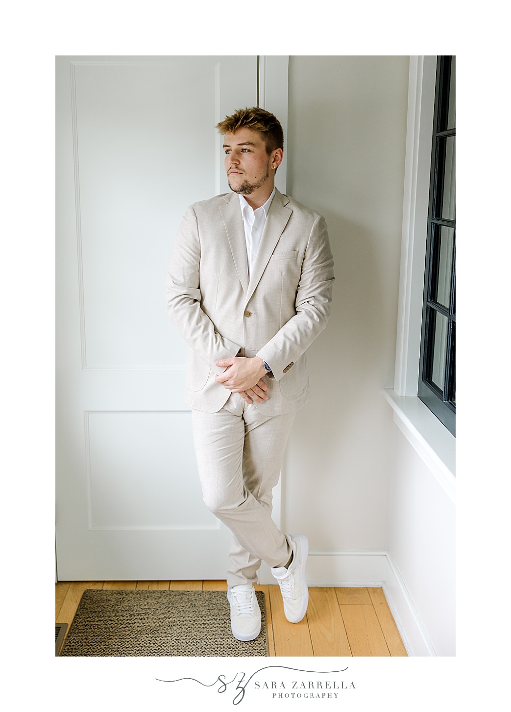 groom leans against white wall in tan suit