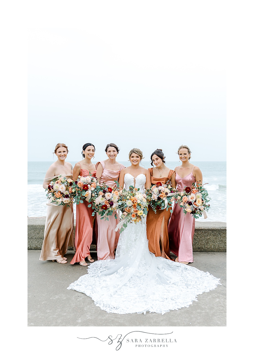 bride and bridesmaids in metallic gowns in various shades of pink pose in front of ocean