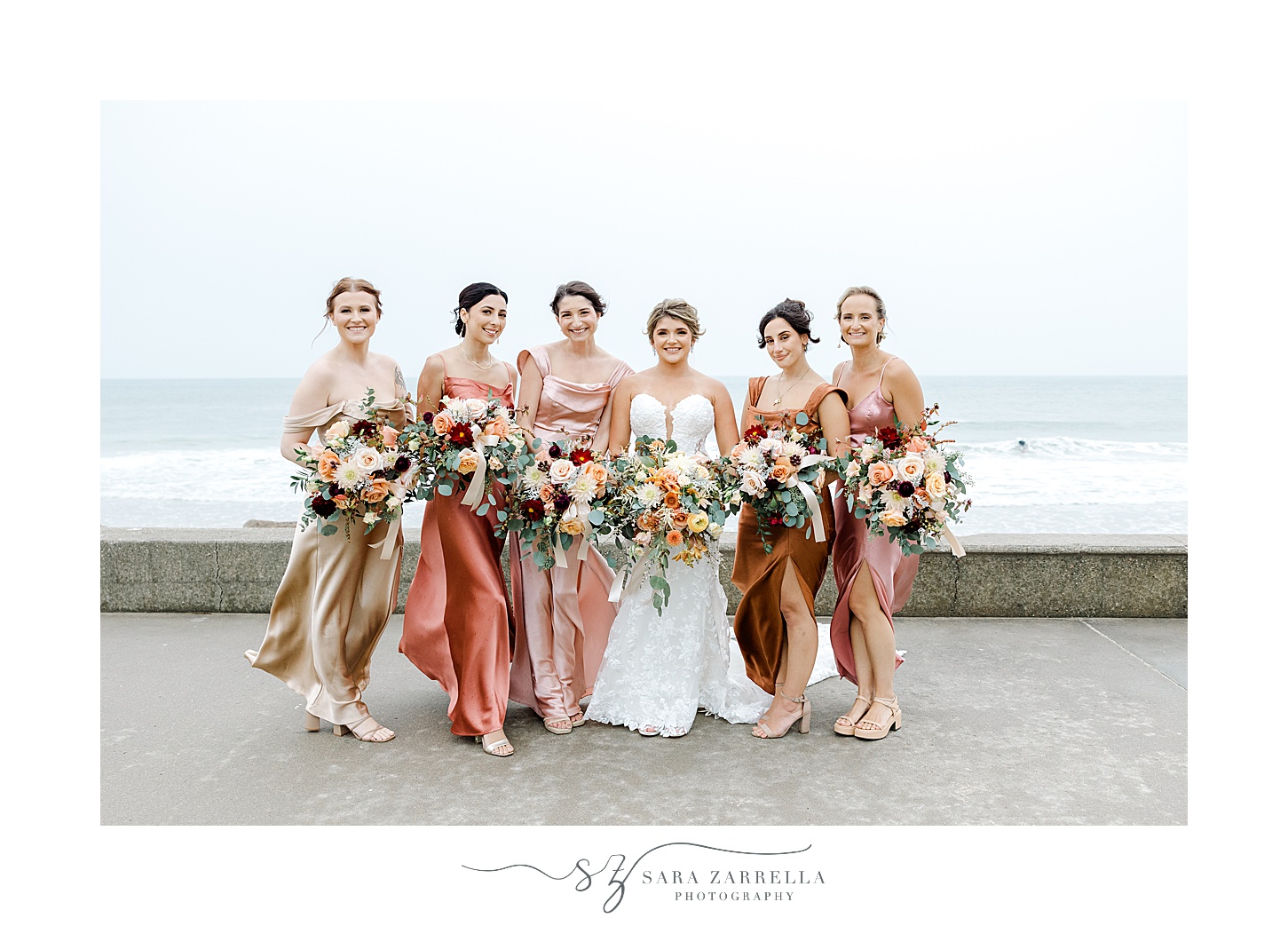 bride and bridesmaids in mismatched pink gowns hug in front of ocean in Westerly RI