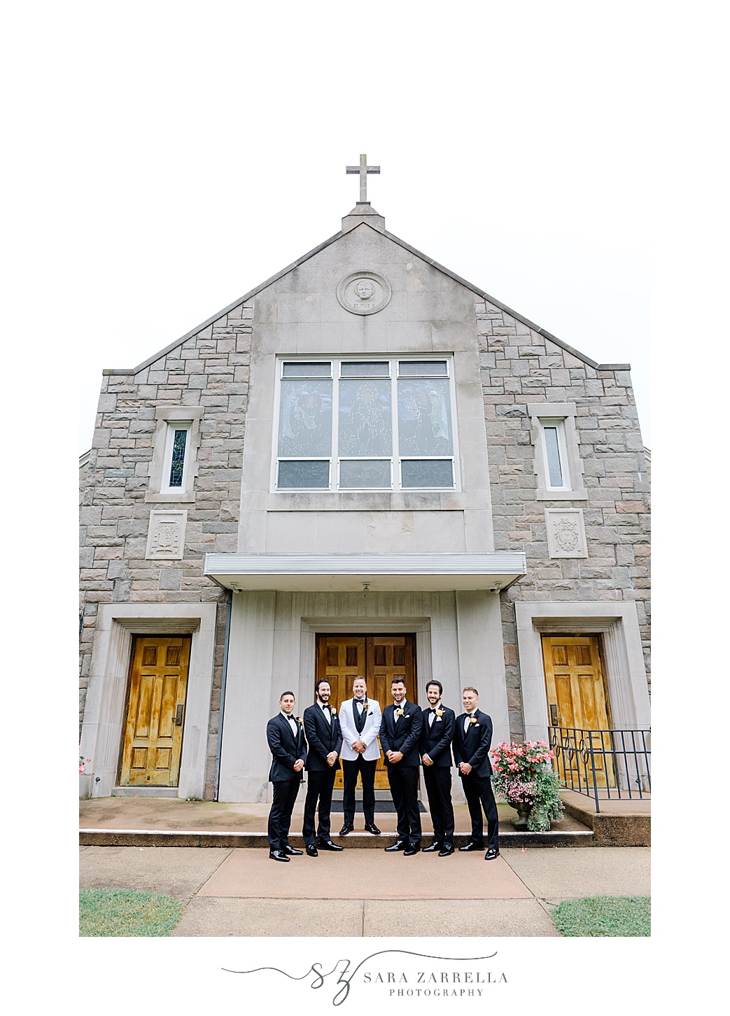 groom stands on front steps of St Pius Church with groomsmen 