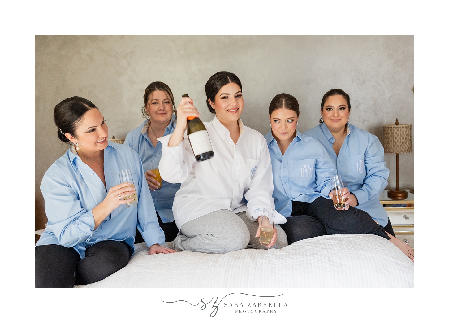 bride sits on bed with bridesmaids in matching blue shirts holding champagne 