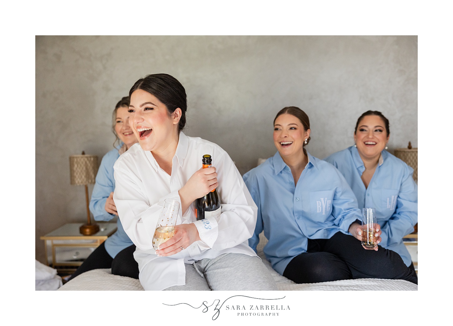 bride laughs holding bottle of champagne on bed with bridesmaids 