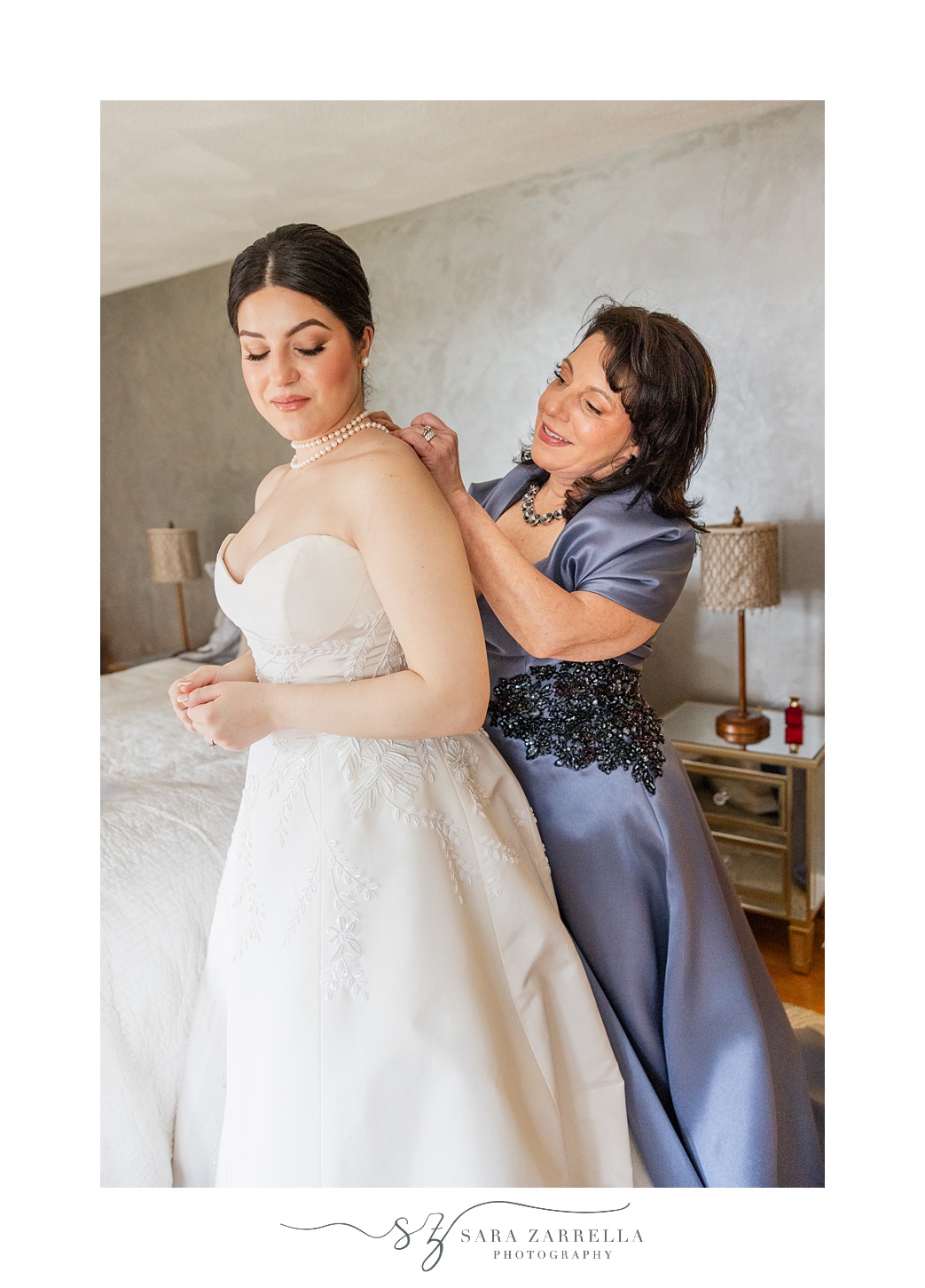 mother in blue dress helps bride into wedding dress and necklace on wedding morning 