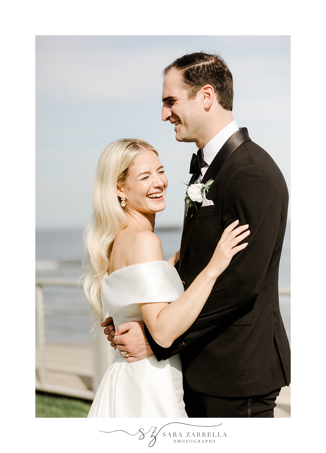 bride holds groom's arms laughing standing on the beach at Narragansett Bay