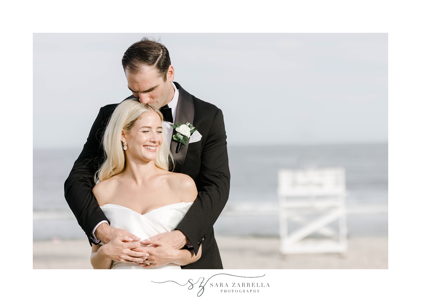 groom hugs bride from behind with Narragansett Bay behind them 