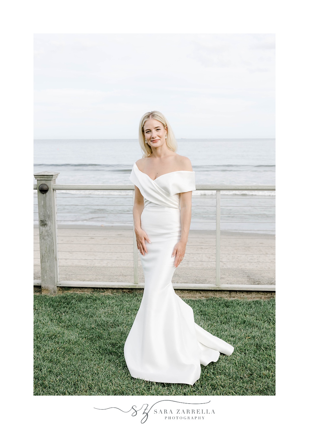 bride in off-the-shoulder ivory gown poses on lawn in front of Narragansett Bay
