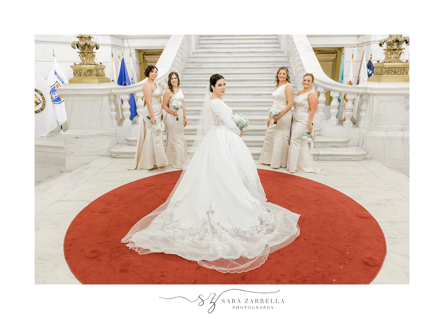 bridesmaids in ivory gowns stand behind bride on red mat inside the Rhode Island State House in Providence RI