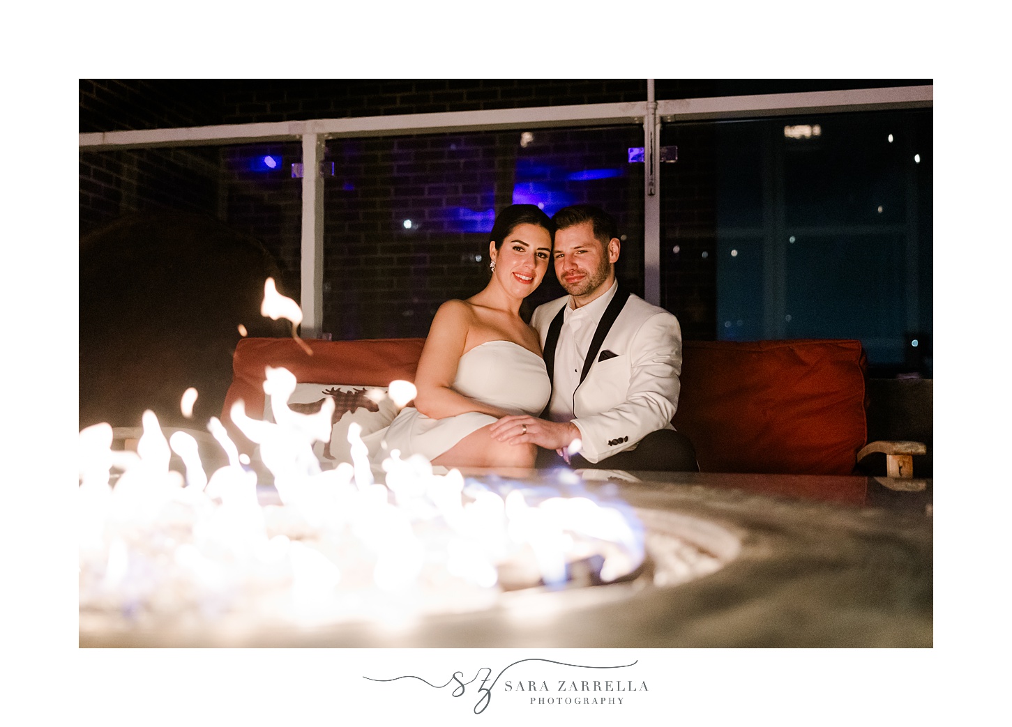groom in white suit jacket with black lapels and bride in short white dress sit by fire pit on rooftop of Providence G