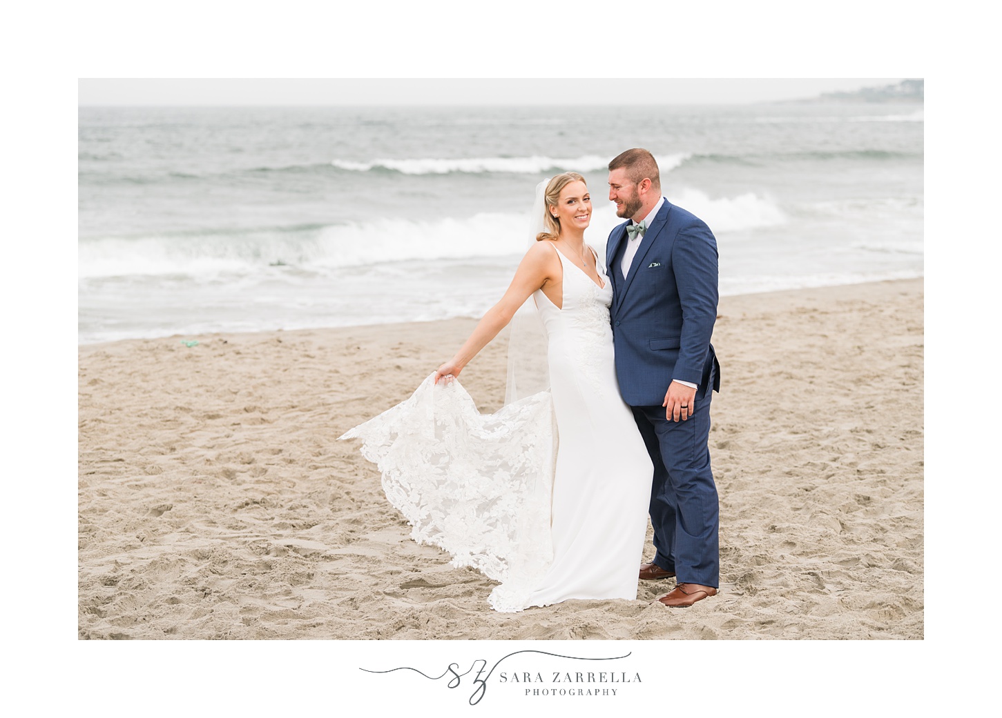 bride holds out skirt of wedding dress standing on beach at Narragansett Bay