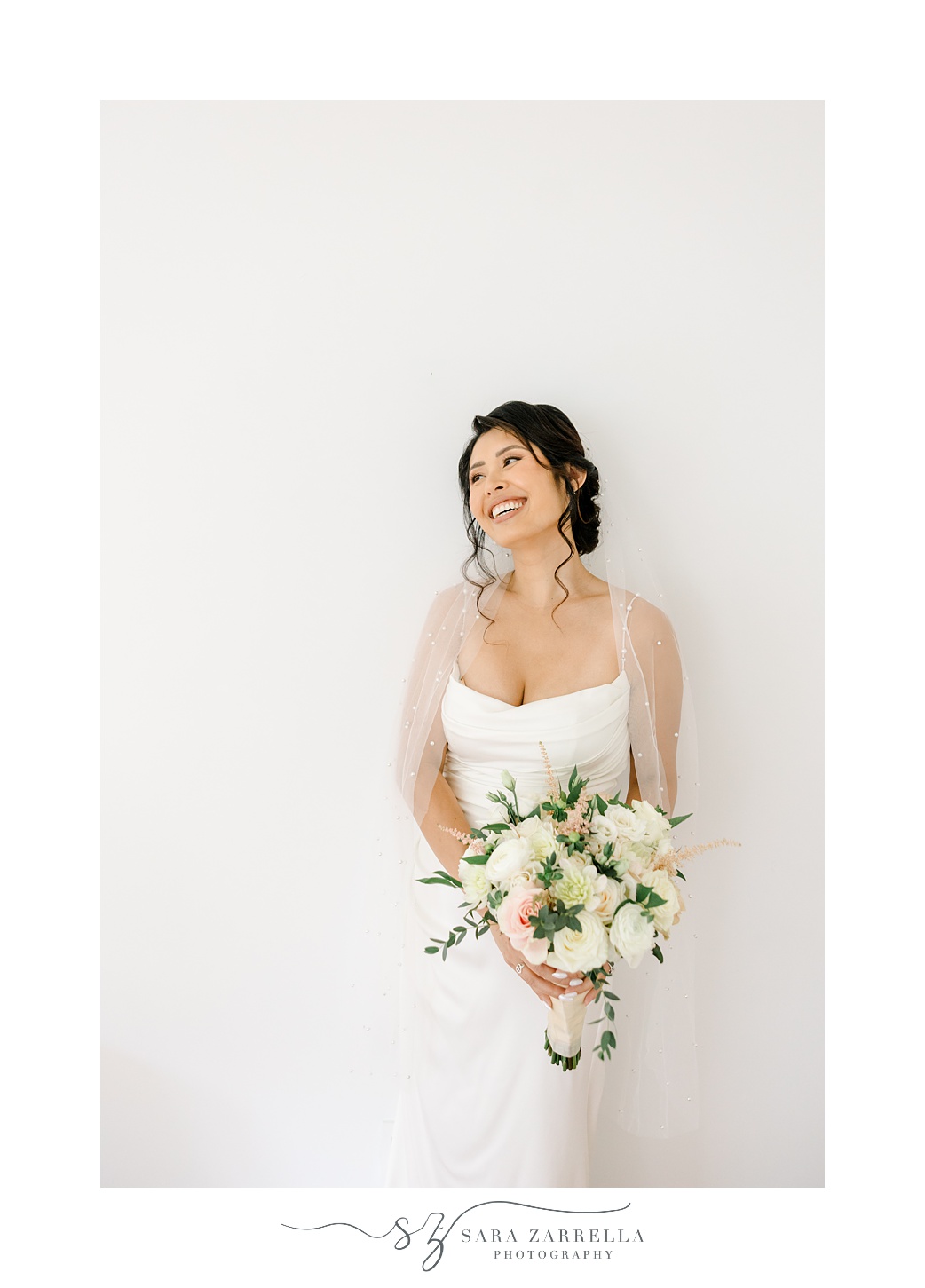 asian bride grins holding bouquet of pink and white flowers against white wall