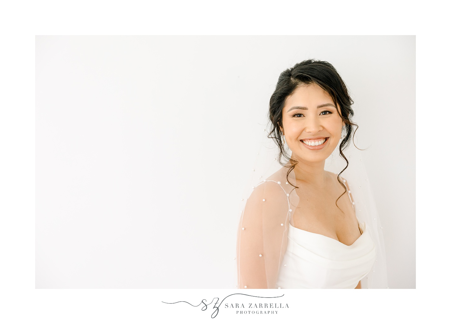 woman smiles in front of white wall in wedding gown with veil around shoulders