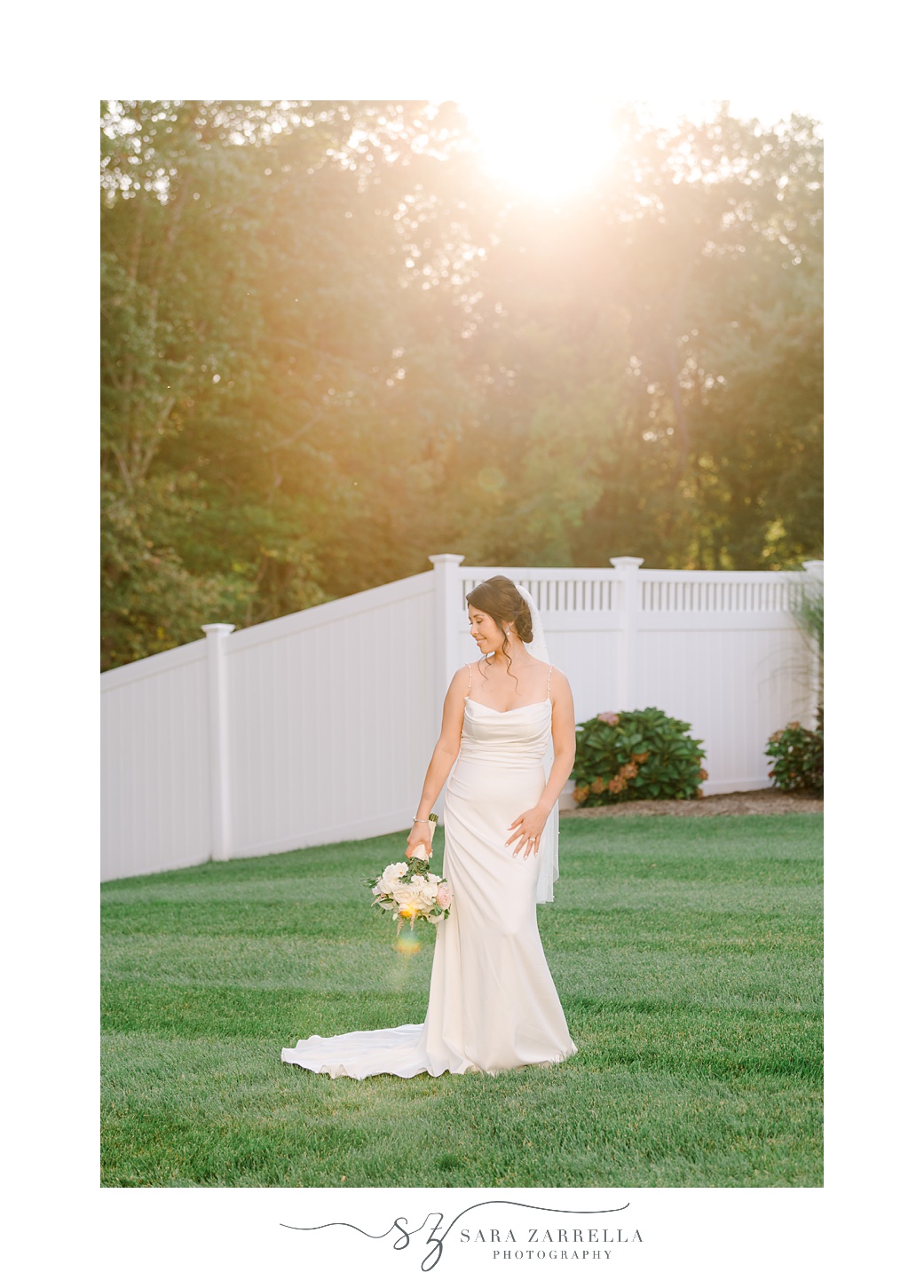 bride stands on lawn at sunset holding bouquet by her hip