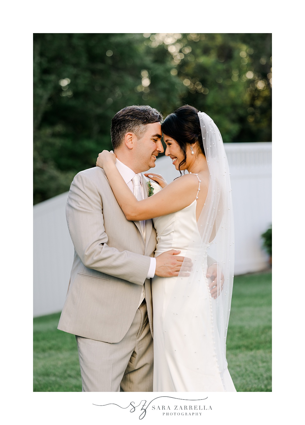 bride and groom hug leaning heads together by white fence in backyard of Cranston RI home