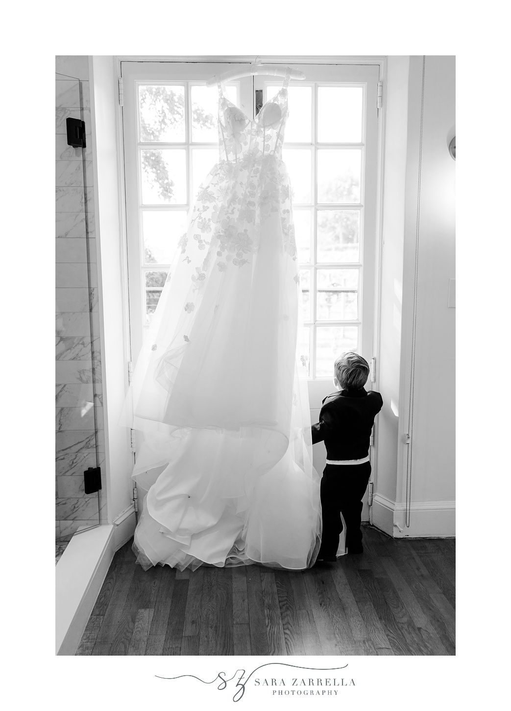 ring bearer looks out window standing by wedding dress hanging in suite at Shepard's Run