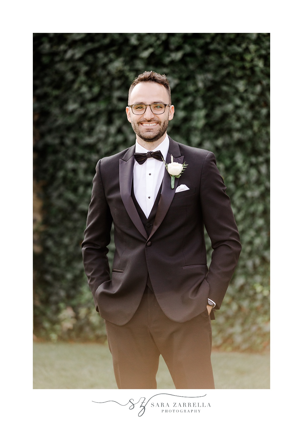 groom stands with hands in pocket of suit in front of ivy covered wall