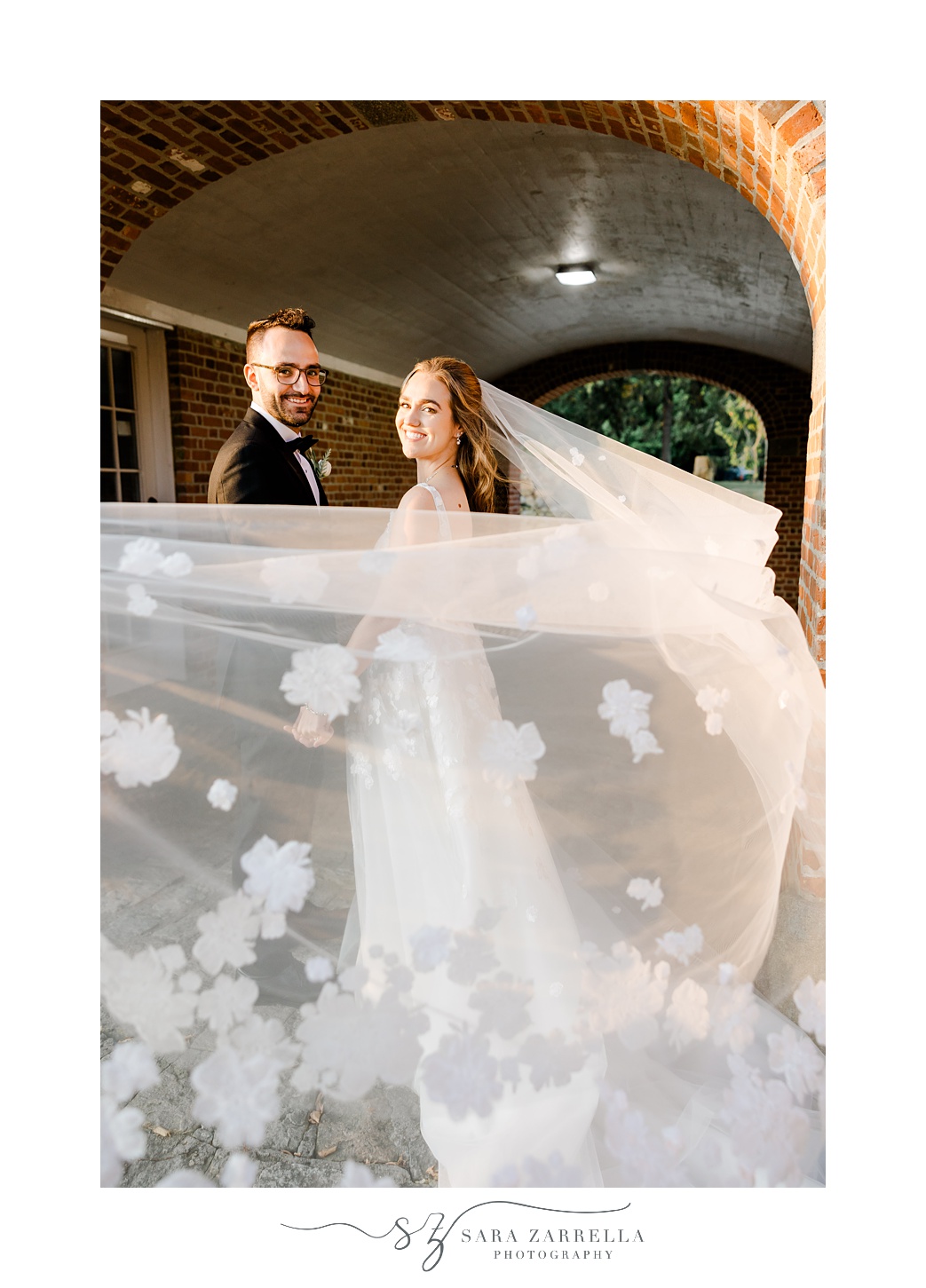 couple looks over shoulders while bride's veil with flower appliqués is pulled by camera