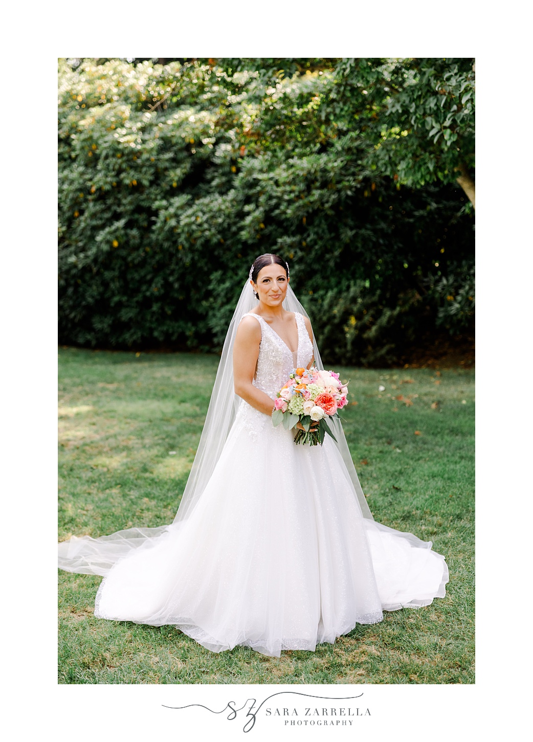 bride stands on lawn holding bouquet with coral and pink flowers