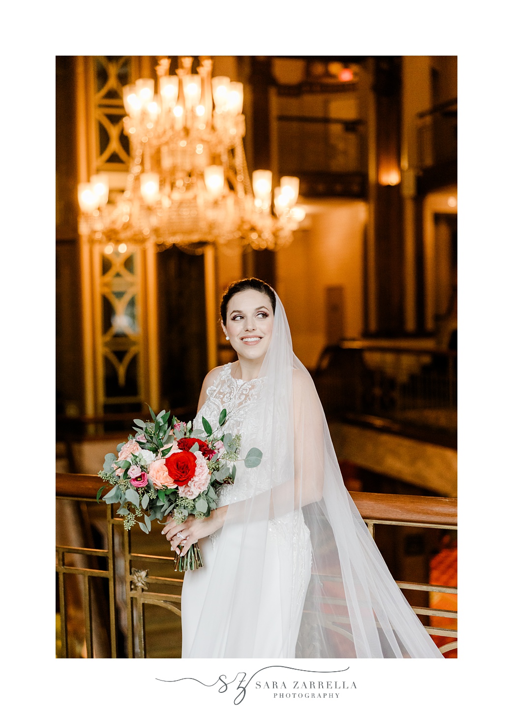 bride stands holding bouquet of red and pink flowers with veil pulled around her arms