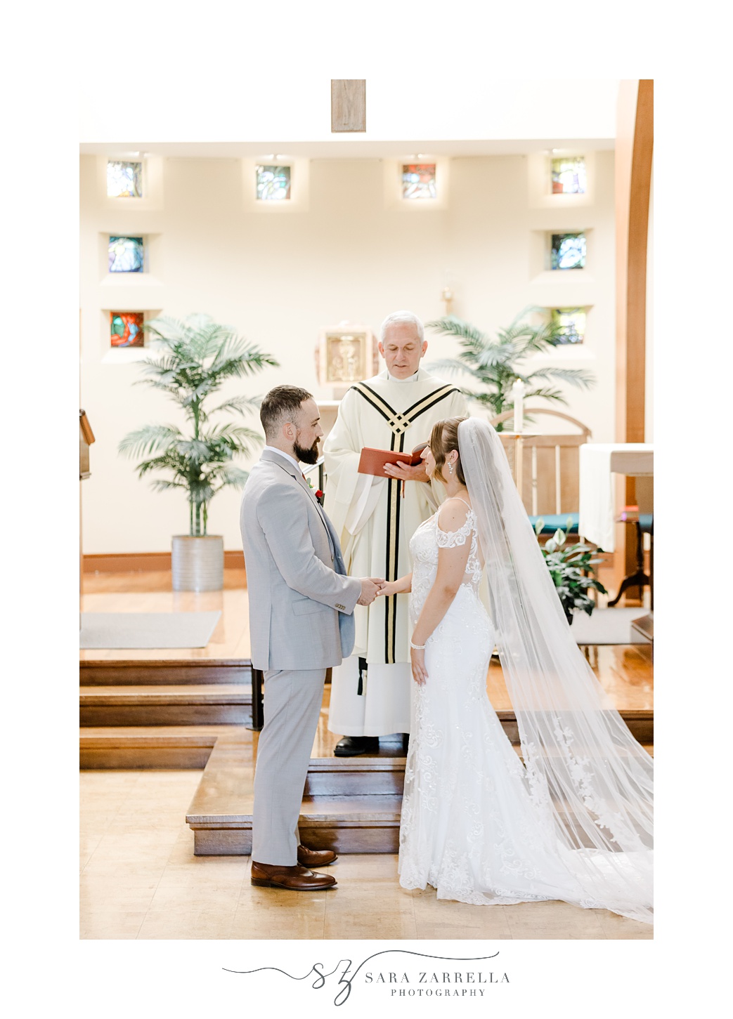 bride and groom hold hands in front of officiant inside Saint John Vianney Parish